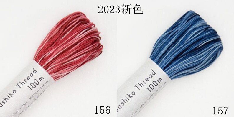 Puede incluir: Dos madejas de hilo Sashiko, una en rojo jaspeado y otra en azul jaspeado, cada una etiquetada como 100m, fabricadas en Jap&oacute;n y marcadas con 2023. La madeja roja est&aacute; marcada como 156 y la azul como 157, ambas de 100% algod&oacute;n.