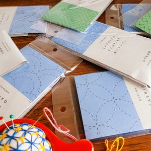 May include: A collection of blue and white Sashiko sampler kits with Japanese text, packaged in clear plastic bags. The kits are arranged on a wooden surface with a small blue and white embroidered pincushion in the foreground.
