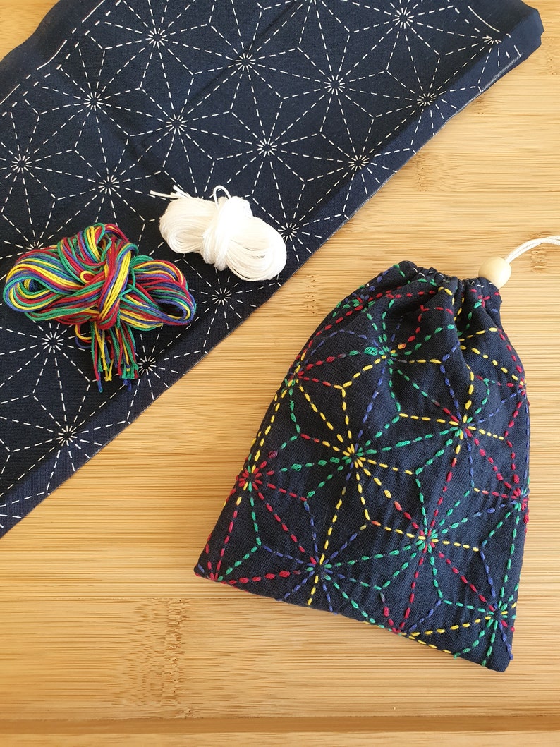 Puede incluir: Una bolsa de tela azul oscuro con un patr&oacute;n geom&eacute;trico de estrellas multicolor, cierre de cord&oacute;n con una cuenta de madera, una pieza de tela azul oscuro a juego con costuras de estrellas blancas, hilo de bordar arco&iacute;ris e hilo blanco.