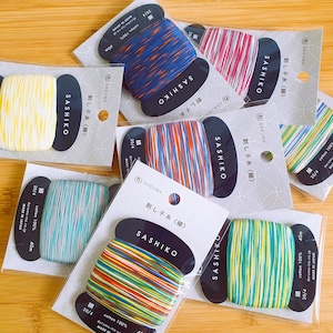 May include: Assortment of colorful Sashiko threads, each packaged on a black and white label. The threads are in various colors, including yellow, blue, red, and green. Each spool is labeled with "Sashiko" and "cotton 100%", and "MADE IN JAPAN".