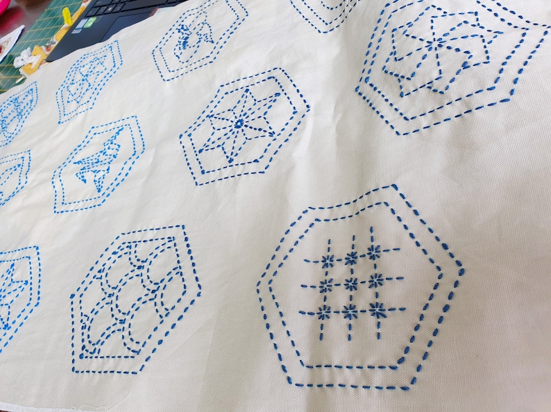 Pu&ograve; includere: Tessuto bianco con cuciture blu in motivi geometrici. I motivi sono esagonali e includono una stella, una griglia e un cerchio.