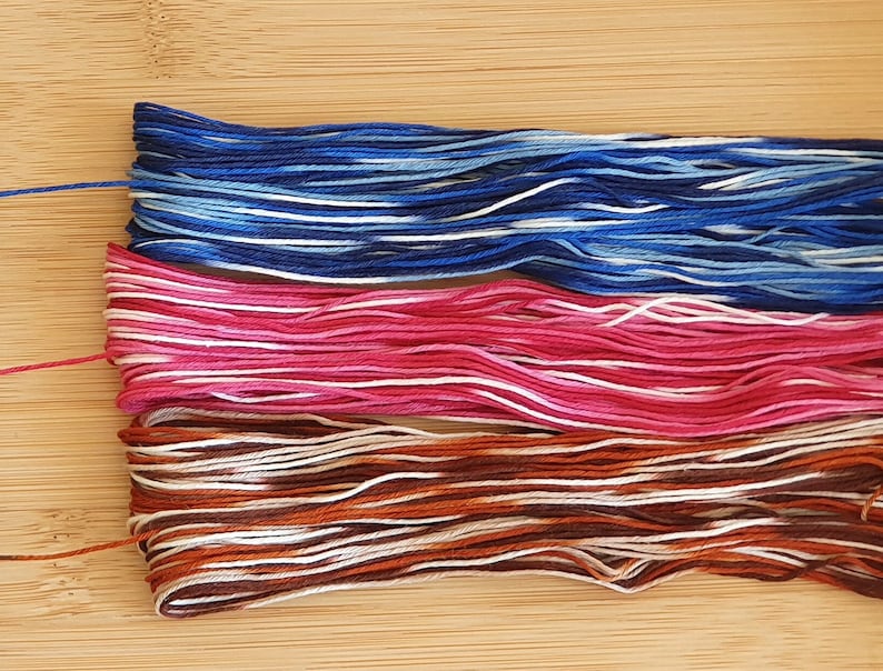 Puede incluir: Tres madejas de hilo de bordar dispuestas horizontalmente. El conjunto superior presenta tonos variados de azul oscuro, azul claro y blanco. El conjunto central muestra rosa, magenta y blanco. El conjunto inferior presenta tonos variados de marr&oacute;n, cobre y blanco.