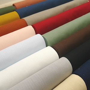 Può includere: Una collezione di rotoli di tessuto in vari colori, tra cui beige, bianco, azzurro, rosa, arancione, giallo, verde, marrone, blu scuro e blu navy. I rotoli di tessuto sono disposti a scalare, con le estremità dei rotoli visibili.