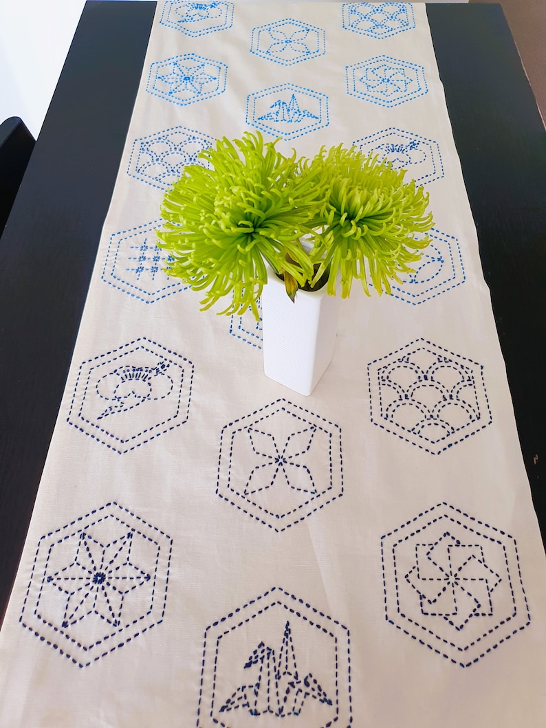 Pu&ograve; includere: Un runner da tavolo bianco con disegni geometrici ricamati in blu. Fiori verdi in un vaso bianco sono centrati sul runner.
