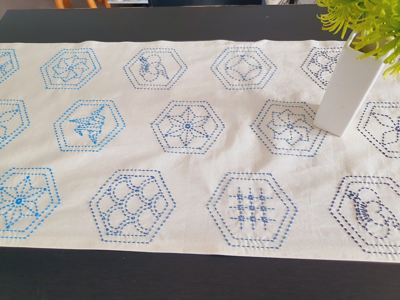 Pu&ograve; includere: Un runner da tavolo in tessuto bianco con disegni esagonali ricamati in blu. I disegni includono forme geometriche, fiori e motivi astratti.