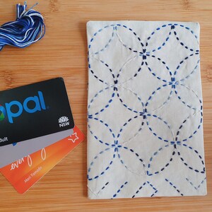 Puede incluir: Un estuche rectangular de tela color hueso con un patr&oacute;n de costura estilo sashiko en azul de c&iacute;rculos entrelazados. Junto al estuche hay un ovillo de hilo de bordar azul y blanco y tres tarjetas de pl&aacute;stico apiladas, incluida una tarjeta Opal.