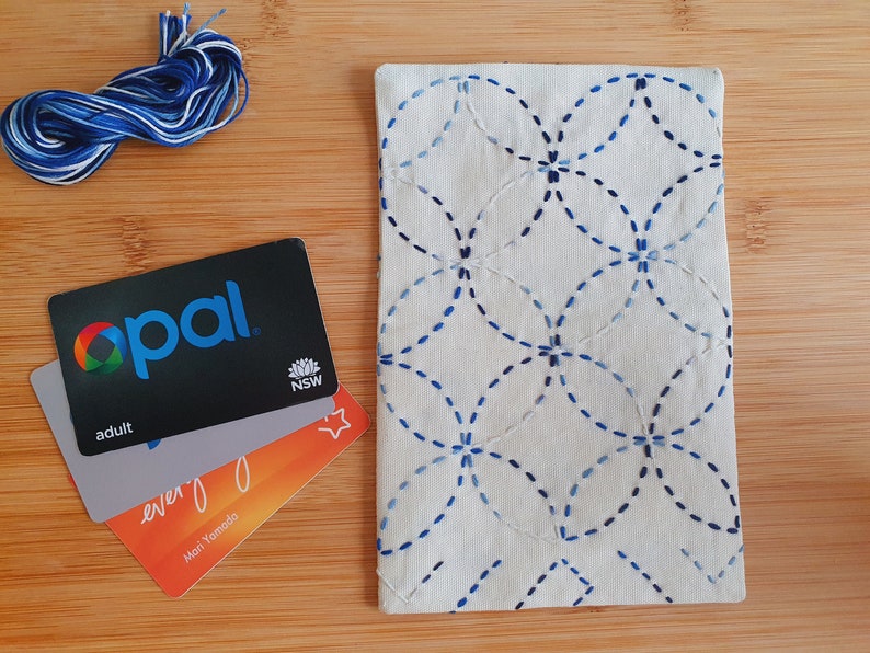 Puede incluir: Un estuche rectangular de tela color hueso con un patr&oacute;n de costura estilo sashiko en azul de c&iacute;rculos entrelazados. Junto al estuche hay un ovillo de hilo de bordar azul y blanco y tres tarjetas de pl&aacute;stico apiladas, incluida una tarjeta Opal.