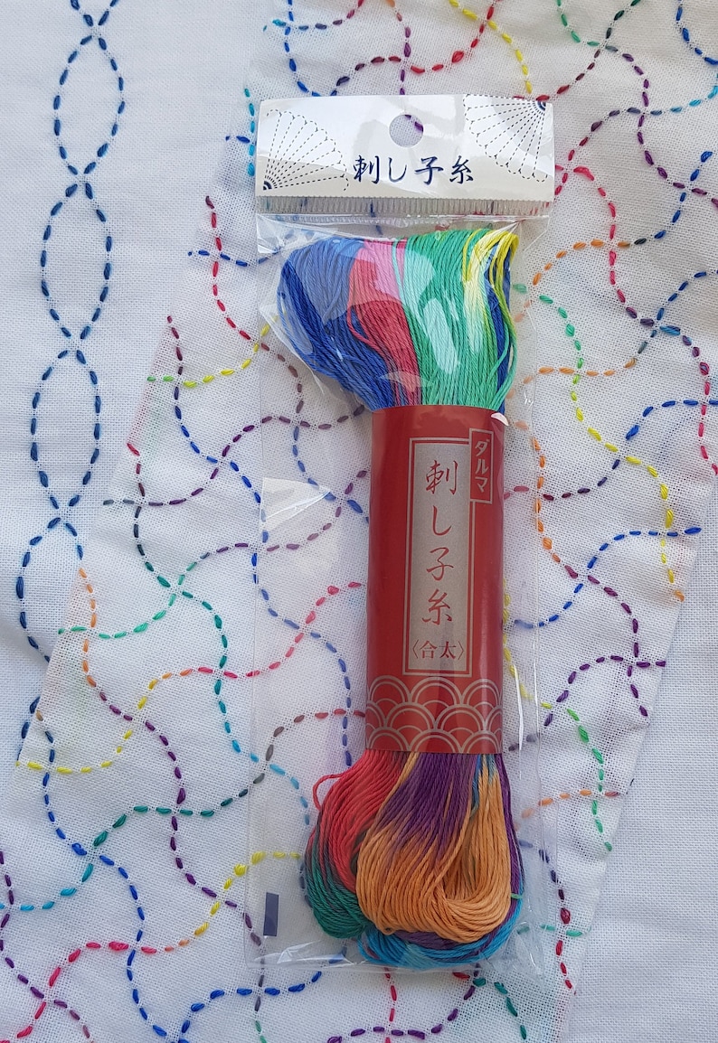 Puede incluir: Una madeja de hilo de bordar multicolor en un paquete de pl&aacute;stico transparente con una etiqueta de papel roja que presenta texto en japon&eacute;s. El hilo muestra un degradado de tonos azules, verdes, amarillos, rosas y morados. El encabezado tambi&eacute;n tiene texto.