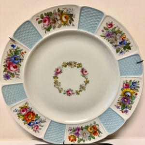 May include: A white porcelain plate with a scalloped edge, featuring a central floral wreath and alternating light blue textured sections and floral bouquets. The flowers are in shades of pink, yellow, blue, and purple.