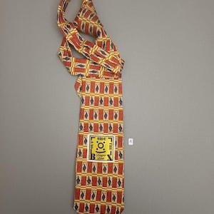 May include: A necktie featuring a repeating geometric pattern of dark blue diamonds on a rust-orange and cream background. A square yellow label is stitched onto the front, displaying the text "OUTSIDE THE BOX Designer Label" with a central icon.