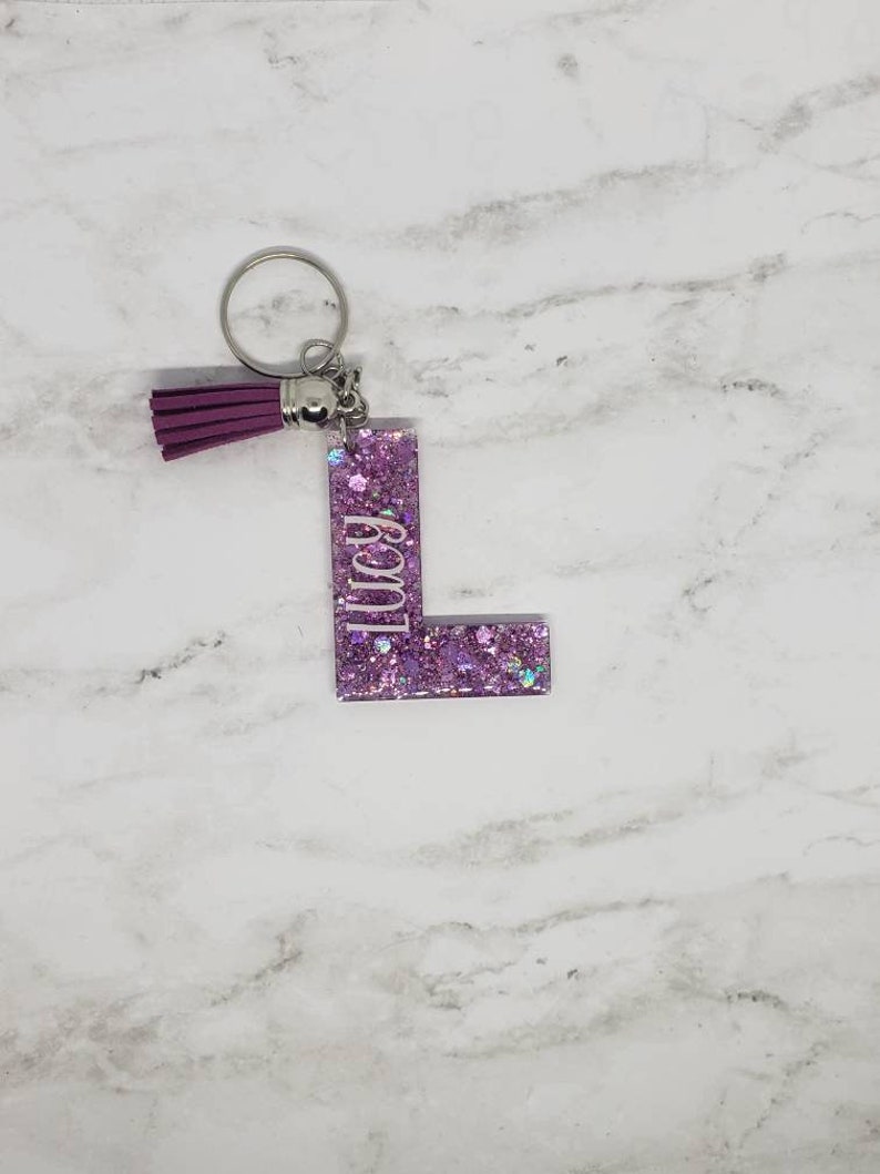 May include: A pink glitter keychain with a purple tassel and the letter "L" with the word "Lucy" written on it.