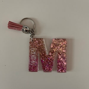 May include: Pink glitter letter M keyring with a pink tassel. The letter M has the name "Mallory" written vertically on the side.