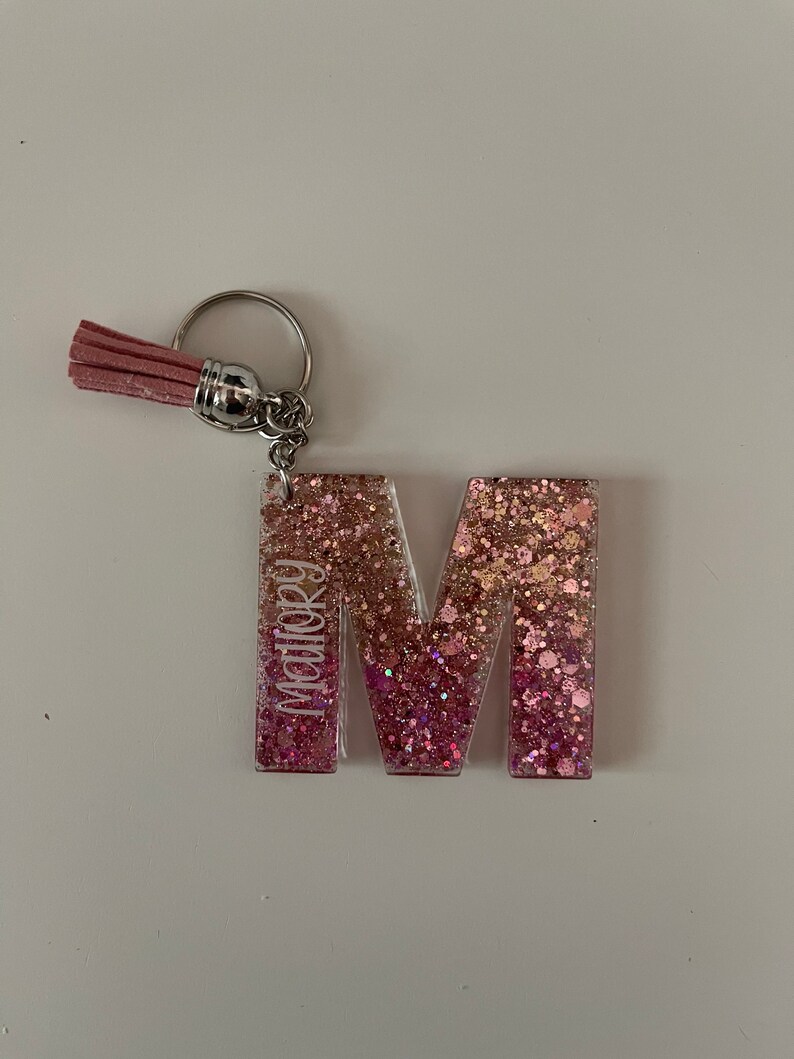 May include: Pink glitter letter M keyring with a pink tassel. The letter M has the name "Mallory" written vertically on the side.
