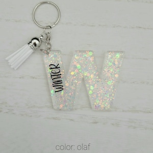 May include: White glitter letter W keychain with a white tassel. The letter has the word "Winter" written on it. The keychain is labeled "color: olaf".