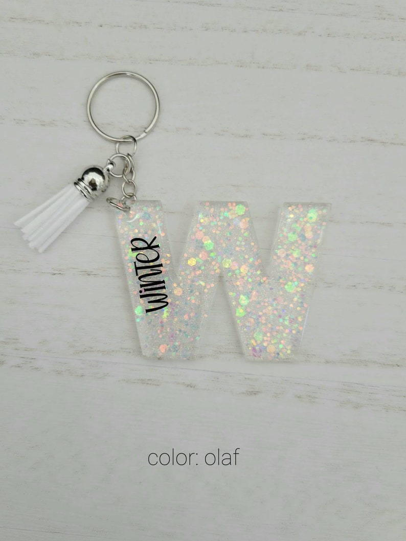 May include: White glitter letter W keychain with a white tassel. The letter has the word "Winter" written on it. The keychain is labeled "color: olaf".