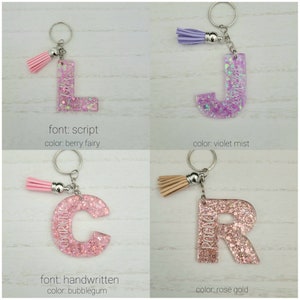 May include: Four personalized glitter keychain charms with different fonts and colors. The charms are in the shape of the letters L, J, C, and R. The letters are filled with glitter and have a tassel attached to the top. The letters are on a white background.