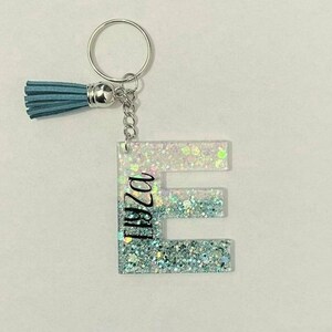 May include: A light blue glitter keychain with a silver key ring and a tassel. The letter "E" is in the centre of the keychain with the name "Eliza" written vertically on the left side of the letter.