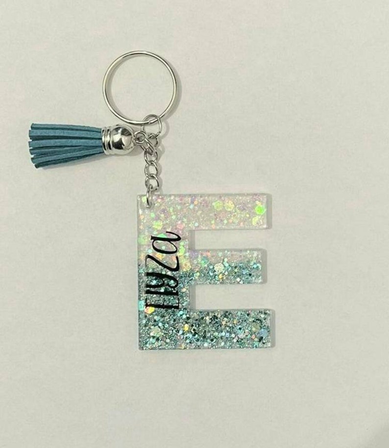 May include: A light blue glitter keychain with a silver key ring and a tassel. The letter "E" is in the centre of the keychain with the name "Eliza" written vertically on the left side of the letter.