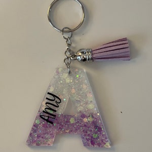 May include: A purple and white glitter resin letter A keyring with a purple tassel. The letter A has the name "Amy" written vertically on the left side.