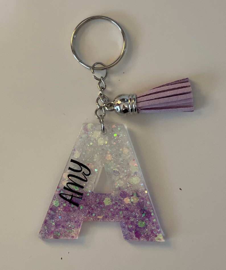 May include: A purple and white glitter resin letter A keyring with a purple tassel. The letter A has the name "Amy" written vertically on the left side.