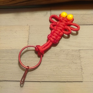 May include: Red and yellow string keychain with a knot design in the shape of a dragonfly. The keychain has a metal ring and a clasp.