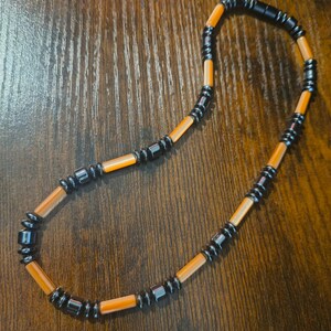 May include: A necklace with alternating orange and black beads. The orange beads are cylindrical, and the black beads are square and clustered. The necklace is displayed on a wooden surface.