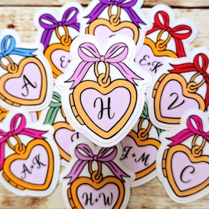 May include: A collection of heart-shaped locket stickers with gold borders and light purple centers. Each locket features a unique letter or monogram in black script and is topped with a colorful bow in shades of pink, red, blue, or purple.