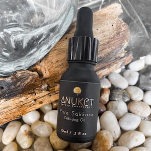 Könnte beinhalten: Eine schwarze Glasflasche mit Tropfer, beschriftet mit "Anuket Luxury Apothecary Pure Sakkara Diffusing Oil 10ml / .3 fl oz".
