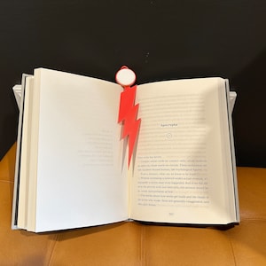 Könnte beinhalten: Ein rotes Blitz-Lesezeichen ist in ein geöffnetes Buch gelegt. Das Buch trägt den Titel "Apocrypha" und der Text auf der Seite lautet: "Jeder Künstler hat seine Helden. Schöpfer, deren Arbeit uns berührt, deren Methoden wir anstreben, deren Welten wir schätzen. Diese außergewöhnlichen Individuen können über das Menschliche hinaus inspirieren, wie mythologische Figuren. Aus der Ferne, was können wir als wahr erkennen? Ohne eine hinter den Kulissen stattfindende, tatsächliche Schöpfung mitzuerleben, ist es unmöglich zu wissen, was wirklich passiert ist. Und wenn wir den Prozess mit eigenen Augen entdecken würden, wäre unser Bericht bestenfalls eine bloße Interpretation. Die Geschichten darüber, wie Werke entstehen und die Rituale der Künstler, die sie geschaffen haben, sind im Allgemeinen übertrieben und oft reine Fiktion."