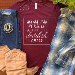 May include: A maroon t-shirt with the text "MAMA HAD HERSELF A LITTLE devilish CHILD" in a white square. The shirt is paired with blue jeans, brown cowboy boots, a blue plaid shirt, and gold hoop earrings. A bracelet is also visible.
