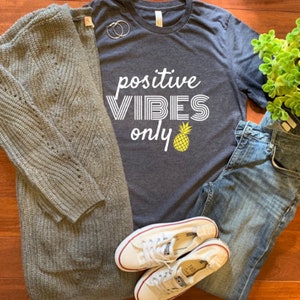 Pode incluir: Um conjunto casual com um cardigan de malha cinza, uma camiseta cinza escuro com o texto "positive VIBES only" e um abacaxi, jeans azuis e tênis de lona brancos. Os itens estão dispostos em uma superfície de madeira.
