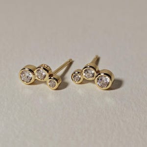 May include: A pair of gold-colored stud earrings, each featuring three clear, round gemstones. The earrings are arranged in a cluster design, with the gemstones varying slightly in size. The studs have a polished finish and are set against a neutral background.