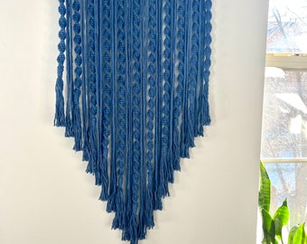 May include: A blue macrame wall hanging with a wooden dowel at the top. The hanging features a series of knots and tassels, creating a geometric pattern.