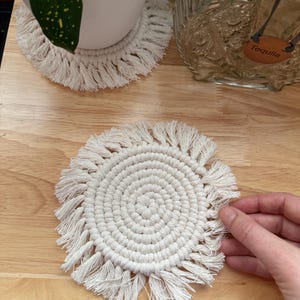 May include: Off-white macrame coasters with a circular design and fringe detail. The coasters are handmade and suitable for use with glasses and mugs. The image shows two coasters, one with a plant on top and one being held by a hand.