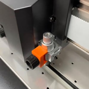 May include: Close-up of a black and silver industrial machine component. The component features a black knob, an orange bracket, and a silver cylindrical piece. The machine has a silver base and a black housing.