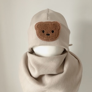 Könnte beinhalten: Eine beige Strickmütze mit einem braunen Teddybär-Applikationen. Die Mütze wird mit einem passenden beigefarbenen Strickschal kombiniert.