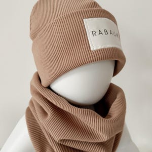 Könnte beinhalten: Eine braune gerippte Strickmütze mit einem weißen Patch, auf dem "RABAUK" steht, und einem passenden braunen gerippten Strick-Halswärmer.