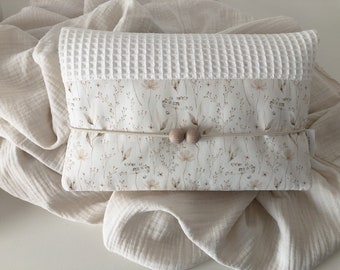 May include: A rectangular, white baby changing pad with a waffle-textured top. The middle section features a floral pattern in beige and brown tones. A beige cord with two wooden beads secures the pad.