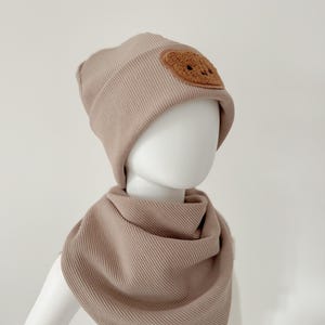Könnte beinhalten: Eine beige gerippte Strickmütze mit einem braunen Filz-Patch, der ein lächelndes Gesicht zeigt. Die Mütze wird mit einem passenden beige gerippten Strickschal kombiniert.
