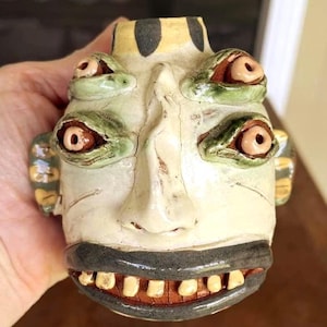 May include: A ceramic sculpture of a head with four protruding eyes, a prominent nose, and a wide mouth featuring visible teeth. The piece is finished in a pale off-white glaze with dark grey accents on the lips and top, and green around the eyes.
