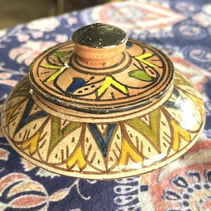 May include: A handcrafted ceramic jar with a lid. The jar is adorned with intricate patterns in yellow, green, blue, and brown. The lid features a black knob and similar decorative elements. This decorative jar is perfect for storing small items.