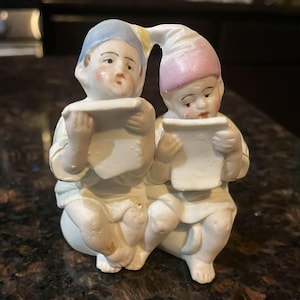 May include: A ceramic figurine depicting two cherubic figures, each holding a scroll. One figure wears a blue hat, the other a pink hat. The figures are dressed in white robes and have rosy cheeks. The figurine is a decorative item.