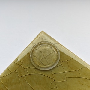May include: A square, green ceramic tile with a crackled glaze. The tile has a clear, round sticker on the bottom.