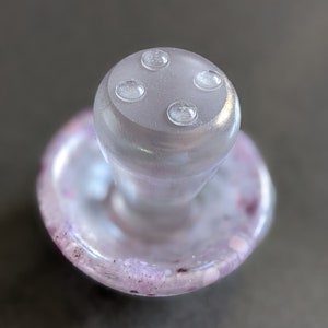 May include: A clear glass bottle stopper with a mushroom-shaped top and four small, clear, raised circles on the top. The base of the stopper is a clear glass disc with a purple and white swirl pattern.