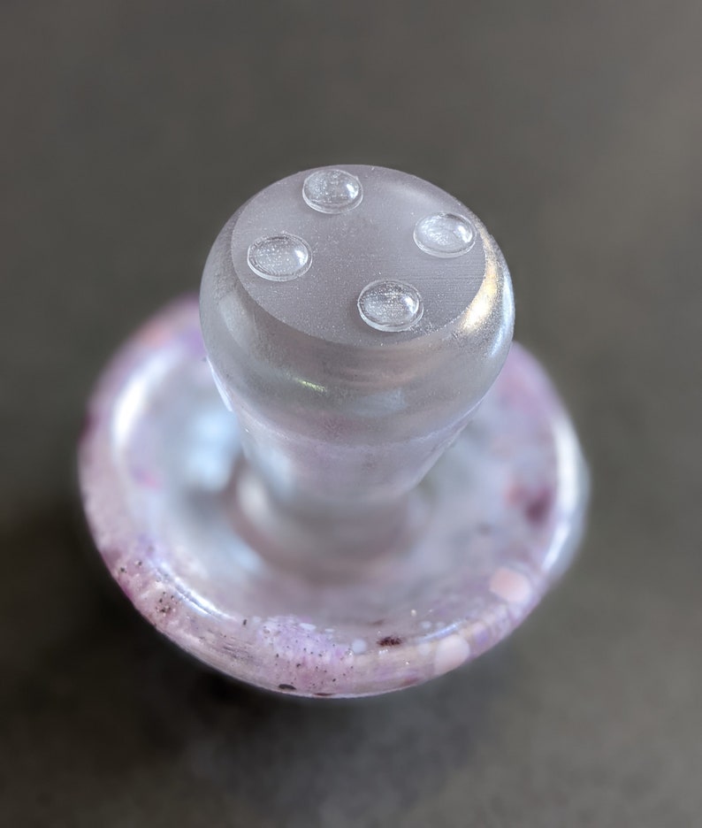 May include: A clear glass bottle stopper with a mushroom-shaped top and four small, clear, raised circles on the top. The base of the stopper is a clear glass disc with a purple and white swirl pattern.
