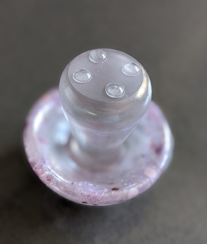 Pu&ograve; includere: Un tappo per bottiglia in vetro trasparente con una parte superiore a forma di fungo e quattro piccoli cerchi chiari in rilievo sulla parte superiore. La base del tappo &egrave; un disco di vetro trasparente con un motivo a spirale viola e bianco.