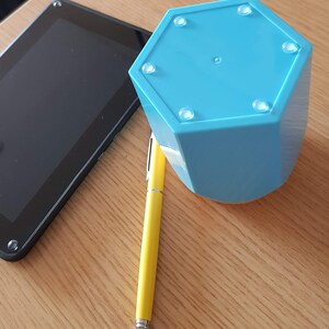 May include: A light blue hexagonal shaped object with six rubber feet on the bottom. The object is sitting on a wooden surface next to a yellow pen and a black tablet.