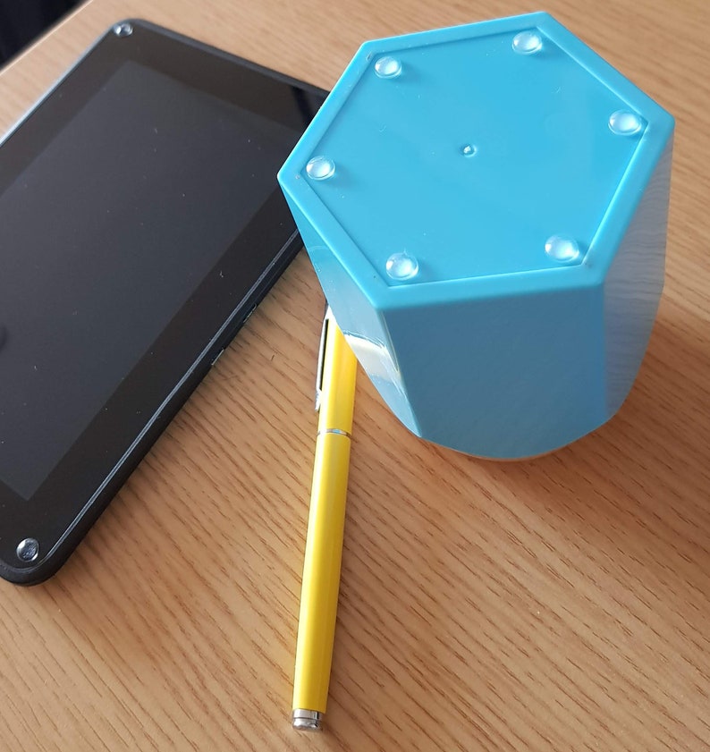 May include: A light blue hexagonal shaped object with six rubber feet on the bottom. The object is sitting on a wooden surface next to a yellow pen and a black tablet.
