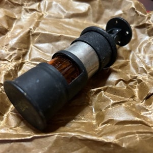 Könnte beinhalten: Ein schwarzer und silberner Flaschenverschluss aus Metall mit einem transparenten Glaseinsatz. Der Verschluss hat eine runde Oberseite und einen kleinen Metallring.