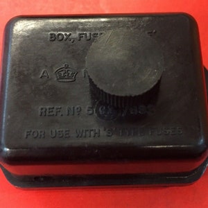 Pode incluir: Caixa de fusíveis preta com botão e orifícios de montagem. A caixa tem texto que diz "BOX, FUSE", "REF. NO 50" e "FOR USE WITH 'S' TYPE FUSES". O fundo é vermelho.